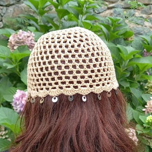 May include: A beige crochet cap featuring an open-work grid pattern and a scalloped bottom edge. The lower border is decorated with a series of small, circular, metallic silver-toned charms that hang evenly spaced around the entire circumference.