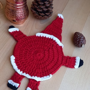 Könnte beinhalten: Ein rot-weißer gehäkelter Weihnachtsmann-Untersetzer. Der Untersetzer ist in Form eines Weihnachtsmanns mit einem runden Körper und zwei Beinen geformt. Der Untersetzer liegt auf einer Holzoberfläche.