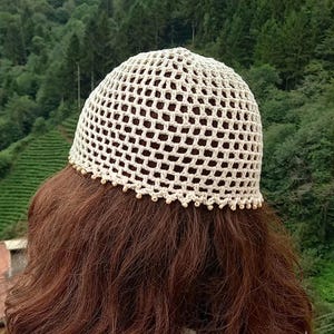 May include: Cream-colored crochet hat with a net-like design. The hat has a rounded crown and a decorative beaded trim along the bottom edge. The hat is worn over brown hair, with a backdrop of green trees.