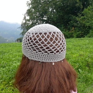 May include: A light gray crochet beanie featuring an openwork net pattern on the crown and a solid band at the base. Small silver-toned star charms are attached at regular intervals along the bottom edge of the hat, resting against hair.