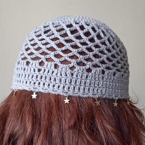 May include: Gray crochet beanie with a star-shaped charm hanging from each side. The beanie has a mesh design and is worn on a person with long brown hair.