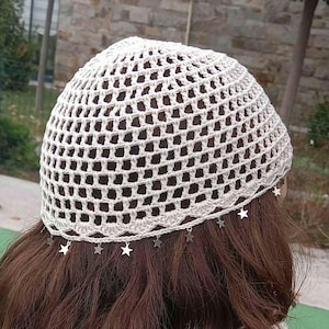May include: A cream-colored crochet beanie featuring an open-work grid pattern. The lower edge of the hat is finished with a scalloped border and is adorned with small, dangling silver-toned star charms spaced evenly around the entire circumference.