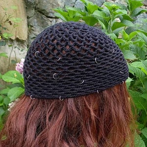 May include: A black crochet beanie featuring a textured, open-weave pattern throughout the crown. The hat is embellished with numerous small, silver-toned metal rings securely attached at various intervals across the dark fabric surface.