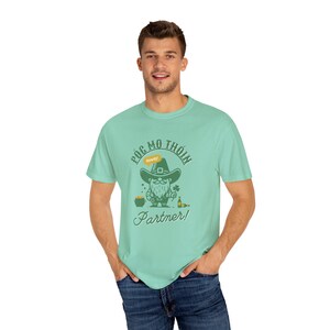 May include: A light green t-shirt with a cartoon leprechaun holding a pot of gold and a beer. The text on the shirt reads "Póg mo thoin Partner!" with a small yellow sign that says "Really?"