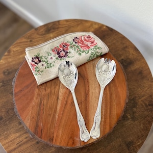 May include: A set of two silver salad servers with ornate pineapple-shaped handles. The servers are on a wooden serving tray with a floral patterned napkin.