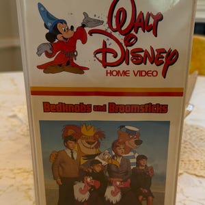 May include: A white Walt Disney Home Video VHS tape case for the movie Bedknobs and Broomsticks. The cover features Mickey Mouse in a blue wizard hat and red robe, with a film still showing three children, a lion, a bear, and two vultures.