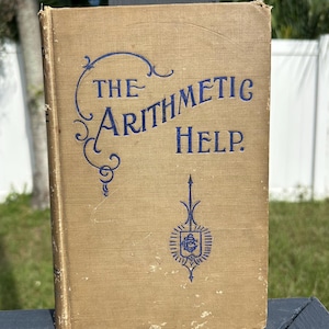 May include: A vintage book titled "THE ARITHMETIC HELP" with blue lettering and decorative flourishes on a tan cover. The book has a worn appearance, suggesting age and use. A blue emblem is featured at the bottom of the cover.
