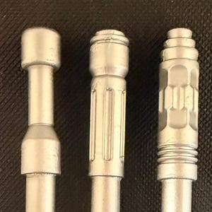 May include: Three silver-colored cylindrical objects arranged vertically. The first has a smooth, tiered top. The second features vertical grooves along its midsection. The third displays a textured, hexagonal pattern with horizontal ridges at the base.