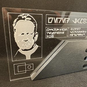 May include: A clear acrylic plaque featuring an etched portrait and text in an alien language. The plaque is supported by a dark grey angled stand with three circular cutouts. The portrait is of a man with a serious expression.