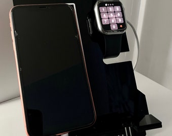 Può includere: Un supporto di ricarica nero per telefono e smartwatch con un simbolo imperiale bianco di Star Wars. Il supporto ha uno slot per un telefono e uno slot circolare per uno smartwatch. Sia il telefono che lo smartwatch sono in carica con cavi bianchi.