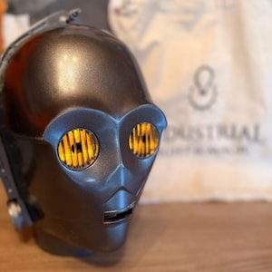 Könnte beinhalten: Eine metallisch bronzefarbene C-3PO-Helmreplik mit leuchtend gelben Augenlinsen. Der Helm hat ein detailliertes Design, einschließlich eines Mundschlitzes und Seitendetails. Im Hintergrund befindet sich eine Tragetasche aus Segeltuch mit einem Logo.