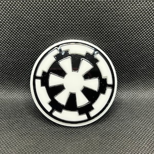 Può includere: Un emblema rotondo in bianco e nero con un design a stella stilizzato. L'emblema ha una finitura lucida ed è posto su uno sfondo nero strutturato. Il design ricorda un impero galattico.