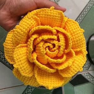 Könnte beinhalten: Eine handgefertigte, leuchtend gelbe Häkelblume mit detaillierter Blütenblattarbeit. Die Blume ist rund und hat eine spiralförmige Mitte mit strukturiertem Aussehen. Der Bastelartikel wird in einer Hand gehalten und zeigt die leuchtende Farbe und das komplizierte Design.
