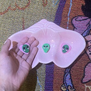 May include: A light pink, three-sectioned ceramic dish shaped like a strawberry. Inside the dish are three small, green alien-shaped charms. Two of the alien charms are attached to pink heart-shaped bases, while one alien charm is plain green.