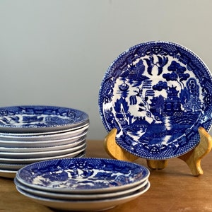 May include: A stack of ten blue and white porcelain saucer plates with a willow pattern design. The plates are stacked on a wooden surface.