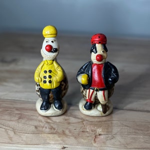 May include: Two small ceramic figurines of clowns. The clown on the left is wearing a yellow jacket with black buttons and a yellow hat. The clown on the right is wearing a red shirt, black pants, and a red hat with black polka dots.