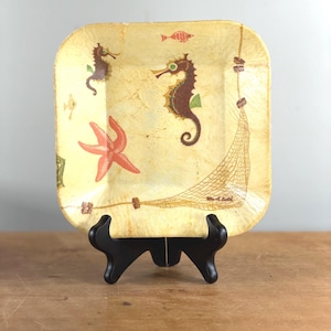 Può includere: Piatto decorativo quadrato a tema vita marina. Il piatto presenta uno sfondo giallo chiaro con cavallucci marini, una stella marina, pesci e un motivo a rete da pesca. Il piatto è esposto su un supporto nero.