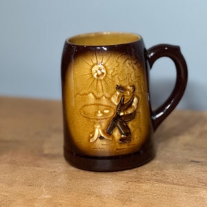 May include: A brown ceramic beer stein with a raised design of a man sitting at a table with a sun in the background.