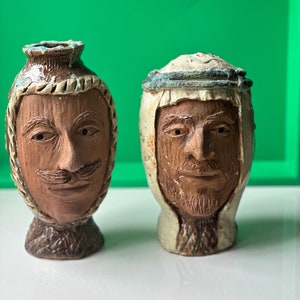 Może przedstawiać: Dwa ceramiczne wazony z rzeźbionymi twarzami. Wazony są brązowe i białe z fakturowaną powierzchnią. Jeden wazon ma brązową brodę, a drugi brązowy wąs.