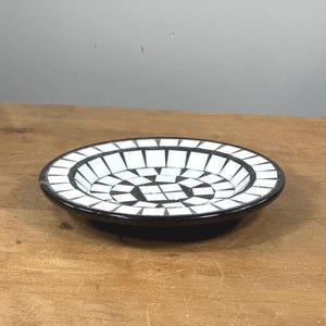Puede incluir: Un plato decorativo pequeño y redondo con un diseño de mosaico. El plato presenta un borde negro y un patrón de baldosas blancas y negras dispuestas radialmente. El plato está sobre una superficie de madera.