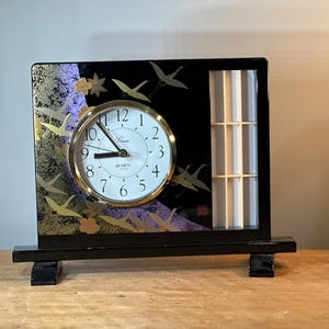 May include: Black and gold tabletop clock with a white face and black hands. The clock has a black frame with a decorative design of cranes and flowers. The clock is on a black stand with two legs.