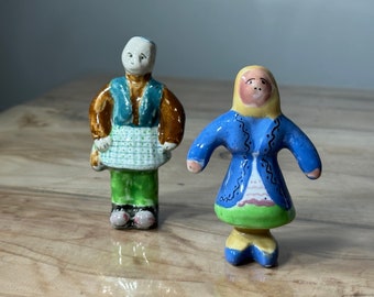 May include: Two ceramic figurines, one with a blue and brown jacket and a green skirt, the other with a blue jacket and a green skirt. Both figurines have painted faces and are standing on a wooden surface.