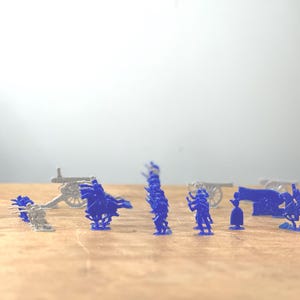 May include: A collection of blue plastic toy soldiers, some on horseback, with cannons and other military equipment. The soldiers are arranged in a battle formation on a wooden surface.