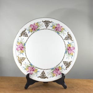 Puede incluir: Plato de cerámica blanco con un diseño floral con flores rosas, amarillas y azules, acentuado con ribetes dorados y elementos decorativos. El plato se exhibe sobre un soporte negro.