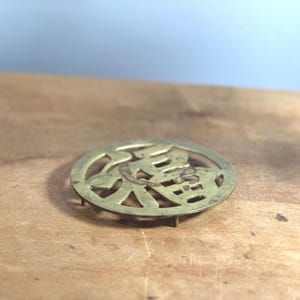 May include: A round, antique brass trivet with a cut-out design featuring Chinese characters. The trivet has small feet and a slightly tarnished finish, suggesting age and use. It rests on a wooden surface.
