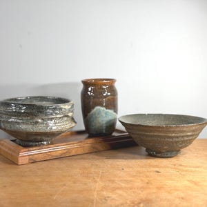 May include: A still life arrangement featuring three ceramic pieces on a wooden tray. The pieces include a textured bowl, a small jar, and a larger bowl. The color palette is earthy, with shades of brown, gray, and blue.