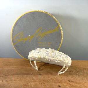 May include: A vintage white beaded headpiece and a round box. The headpiece features a delicate design with small beads and rhinestones. The box is grey with gold lettering that reads "Sharry Originals".