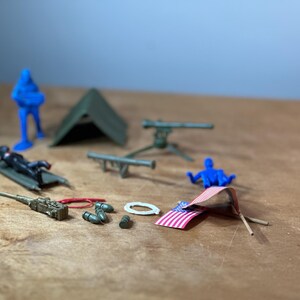 Puede incluir: Una colección de figuras y equipos militares de juguete, que incluyen una tienda de campaña verde, un soldado azul, una ametralladora, una camilla y una pequeña bandera estadounidense.