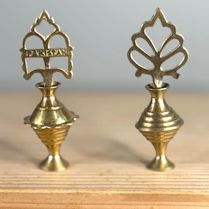 Könnte beinhalten: Zwei verzierte Messingbehälter. Einer hat das Wort "PAKISTAN" in einem dekorativen Rahmen. Der andere hat ein blattähnliches Design. Beide haben einen bauchigen Sockel und stehen auf einer Holzoberfläche.