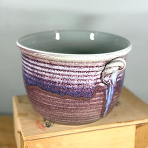 Może przedstawiać: Ceramiczna miska o okrągłym kształcie i szerokim otworze. Miska ma teksturowany, poziomy wzór w paski w odcieniach fioletu, brązu i bieli. Ma szare wnętrze i mały uchwyt z boku. Miska stoi na drewnianej skrzyni.