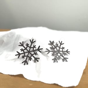 Peut inclure: Deux boucles d'oreilles en forme de flocon de neige gris foncé. Chaque boucle d'oreille a six pointes avec de petites extensions à l'extrémité de chaque pointe. Les boucles d'oreilles sont présentées sur un tissu blanc.