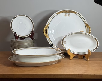 May include: A set of white porcelain dishes with gold trim. The set includes a large platter, a small oval dish, a small round dish, and a set of stacked plates. The dishes are decorated with a gold band and a gold monogram.