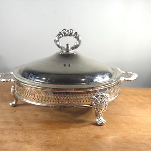 May include: Silver-plated serving dish with a glass lid and ornate handles. The dish features a decorative pierced design around the base and rests on three detailed feet. The lid has a decorative handle.