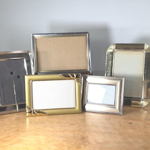 May include: Five assorted picture frames in silver and gold tones. The frames vary in size and shape, with rectangular and square designs. Some frames have a simple, sleek design, while others feature decorative details.