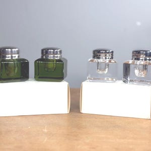 May include: Four square glass inkwells with silver-toned metal caps, arranged in two pairs. Two inkwells are made of translucent green glass, and two are made of clear glass. Each pair sits atop a plain white rectangular cardboard box.
