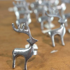May include: A silver-toned metal napkin ring featuring a stylized reindeer silhouette with textured detailing on the body and antlers. The reindeer stands upright with a circular ring base attached to its side, presented in a group of multiple units.
