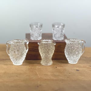Könnte beinhalten: Eine Sammlung klarer, vintage Glas Sahnekännchen und Schnapsgläser. Die Sahnekännchen haben Griffe und ein Sternmuster. Die Schnapsgläser sind zylindrisch mit einem strukturierten Design. Sie sind auf einer Holzoberfläche angeordnet.