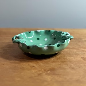 May include: A small, green ceramic bowl with white and black polka dots. The bowl has a ruffled edge and two small handles.