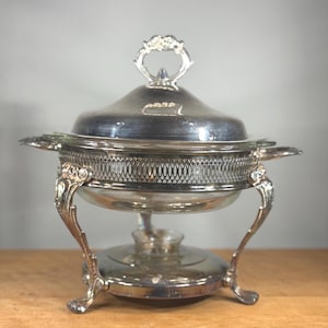Op de afbeelding: Zilveren chafing dish met een gewelfde deksel en sierlijke handvatten. De schaal heeft een glazen inzetstuk en decoratieve filigraan rond de basis. De poten zijn gebogen en gedetailleerd, en de handgreep op het deksel is bloemig.