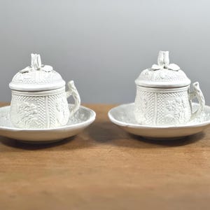 Puede incluir: Dos tazas de cerámica blanca con tapas y platillos. Las tazas tienen un patrón floral detallado y un asa. Las tapas están rematadas con un adorno en forma de flor. Las tazas se asientan sobre platillos a juego, todo en un acabado blanco mate.