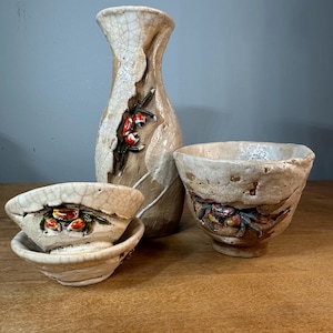 May include: A set of three ceramic sake cups and a bottle, all with a crackled glaze and decorative floral and crab motifs. The cups are stacked on top of each other, and the bottle is standing upright.