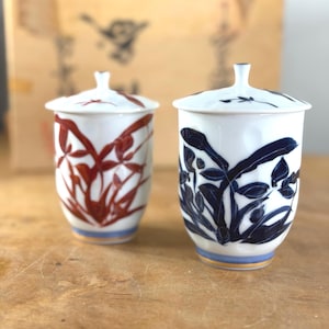 May include: Two white ceramic tea cups with lids. One cup features a red floral design, the other a blue floral design. Both have a blue and gold band at the base. The lids have a small handle.