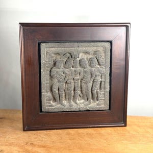 May include: A square dark brown wooden frame containing a gray stone relief carving. The carving depicts four standing figures in a row, rendered in a textured, weathered style with arched architectural details visible above the figures' heads.