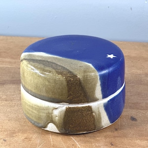 Peut inclure: Un pot en céramique avec un couvercle. Le pot est cylindrique avec un dessus bleu et une étoile blanche. La section du milieu est beige et marron, avec une bande blanche séparant le haut et le bas.
