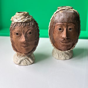 Puede incluir: Dos jarrones de cerámica con forma de cabeza humana con caras marrones y cintas blancas. Los jarrones están sobre una superficie blanca.