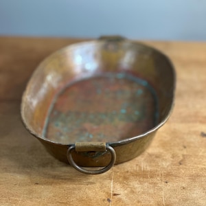 Puede incluir: Bandeja ovalada de metal color cobre con dos asas de anillo. La bandeja tiene un aspecto ligeramente envejecido con zonas de oxidación verde y roja, lo que sugiere antigüedad. Las asas están fijadas con soportes rectangulares. La bandeja está sobre una superficie de madera.